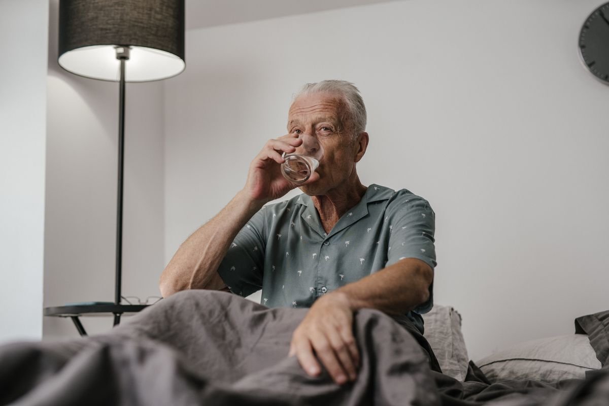 Senior man with gray hair drinking a glass of water in a bedroom setting
