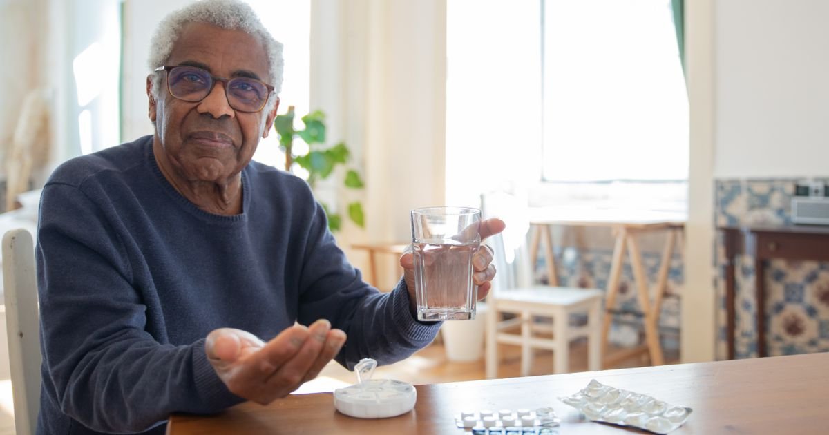 Older man sitting at a table holding a glass of water and medication