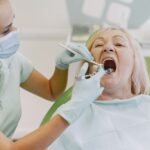 A dentist wearing a mask and gloves examining a senior woman's teeth and gums in a dental clinic