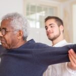 A senior man is assisted by a caregiver with a shoulder stretching exercise indoors