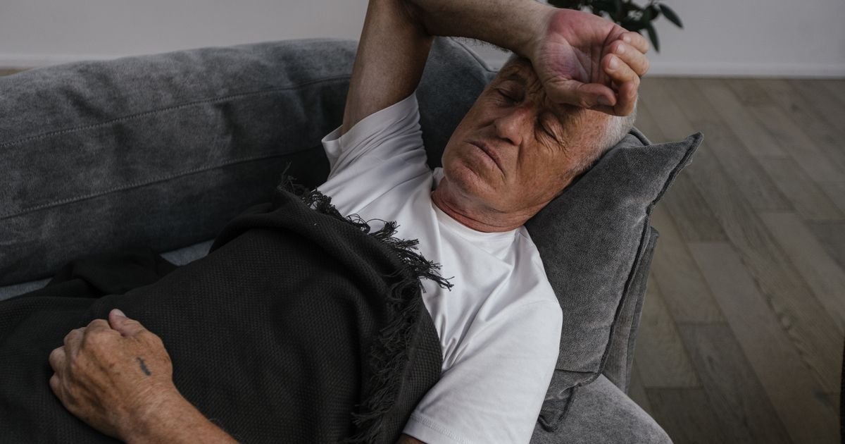 Older man resting on a sofa with his hand on his forehead, looking fatigued
