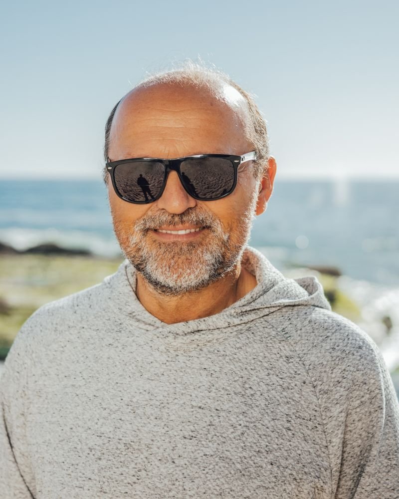 Smiling senior man with a beard and sunglasses standing by the coast, looking healthy and vibrant