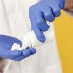 Pharmacist in a white coat wearing blue gloves pouring white pills from a bottle into a hand
