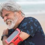 Senior man with gray hair and beard grimacing and holding his shoulder in pain