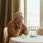 Senior man sitting thoughtfully at a table by a window, with glasses and medication nearby