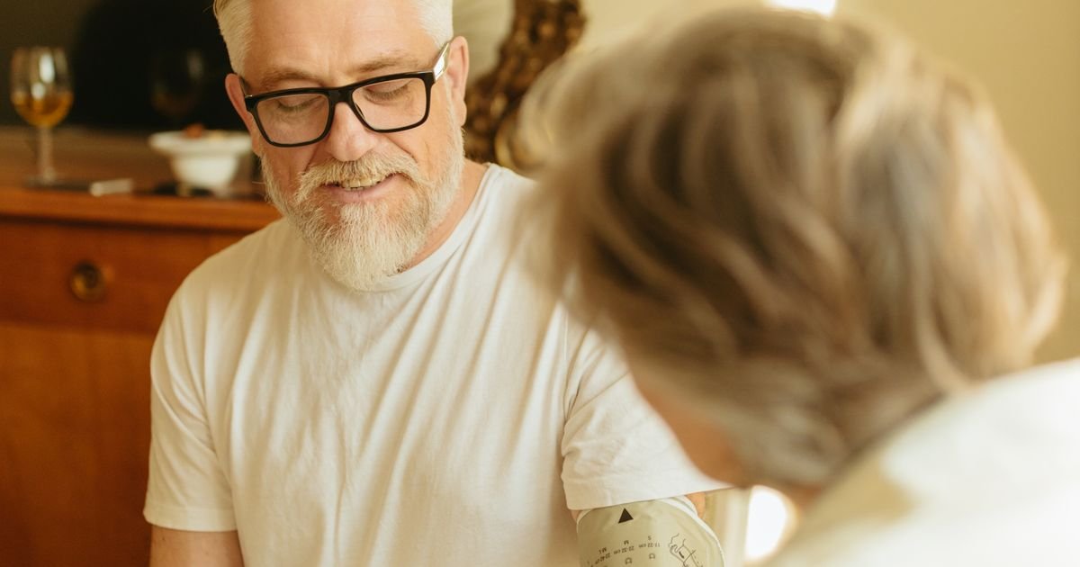 Senior man with gray hair and glasses checking his blood pressure with a digital monitor at home.