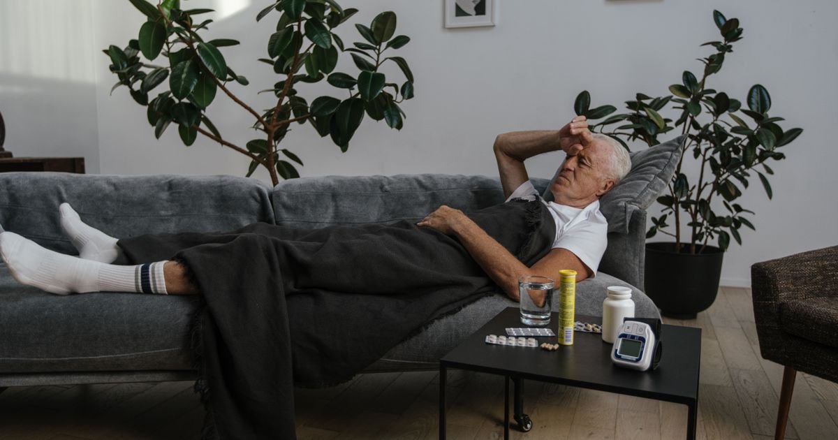 Senior man resting on a couch at home with medication on the table nearby, looking unwell.